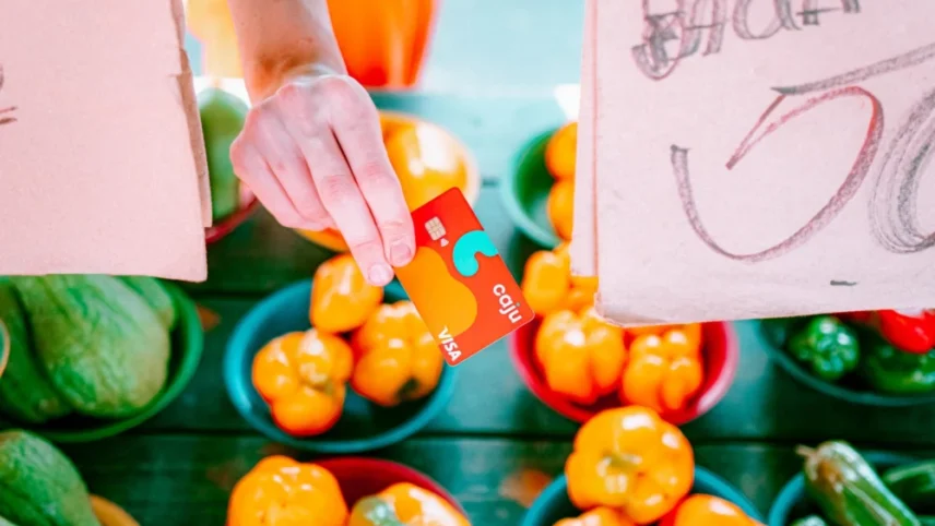 Mão segurando o cartão Caju Benefícios próximo a frutas diversas distribuídas em uma mesa.